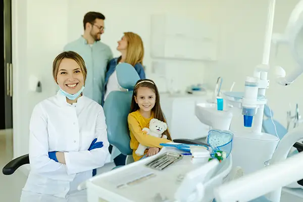 A young girl sitting in dentist chair with teddy bear in arms, smiling with parents and dentist after a dental visit.